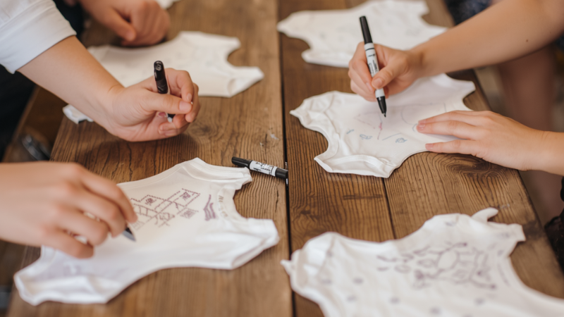 A DIY onesie decorating station at a baby shower, guests hands drawing creative patterns on white baby bodysuits, non-toxic fabric markers on a wooden table, artistic and heartwarming atmosphere, candid shot, lifestyle photography.