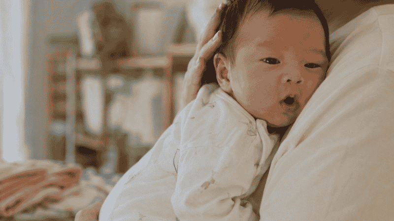 A newborn baby hiccupping after feeding while being held upright against a parent's chest