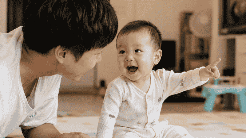 A baby around 12 months old pointing at something and vocalizing their first words to a parent