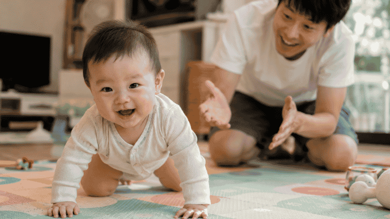 A baby crawling on all fours across a soft play mat toward a parent who is smiling and encouraging them