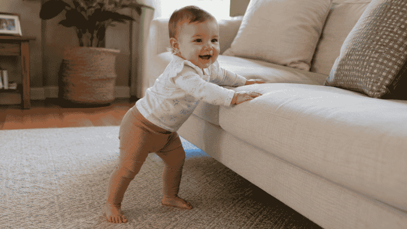 A baby cruising sideways along a couch while holding on with both hands, practicing pre-walking movement