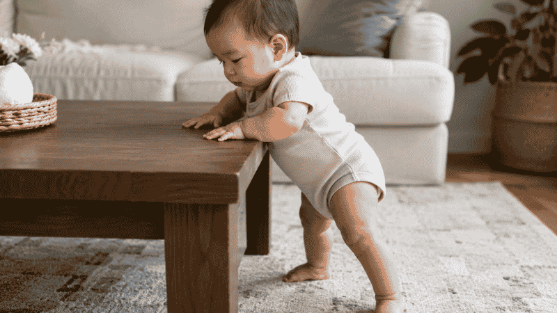 A baby around 9 months old pulling themselves up to stand by holding onto a low wooden coffee table