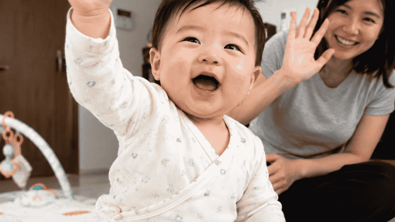 A baby around 8 months old waving one hand in a bye-bye gesture during a social interaction
