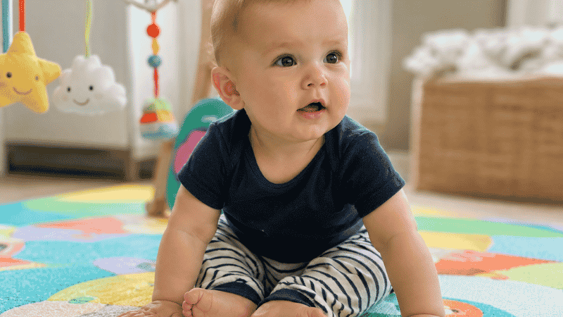 A baby around 6 months old sitting up independently on a soft play mat, looking curious and alert