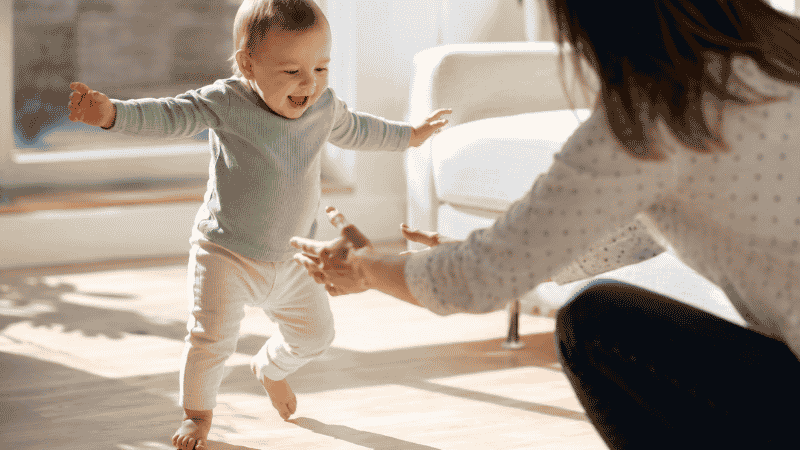 A baby taking their first independent steps across a living room floor with arms outstretched and a proud expression
